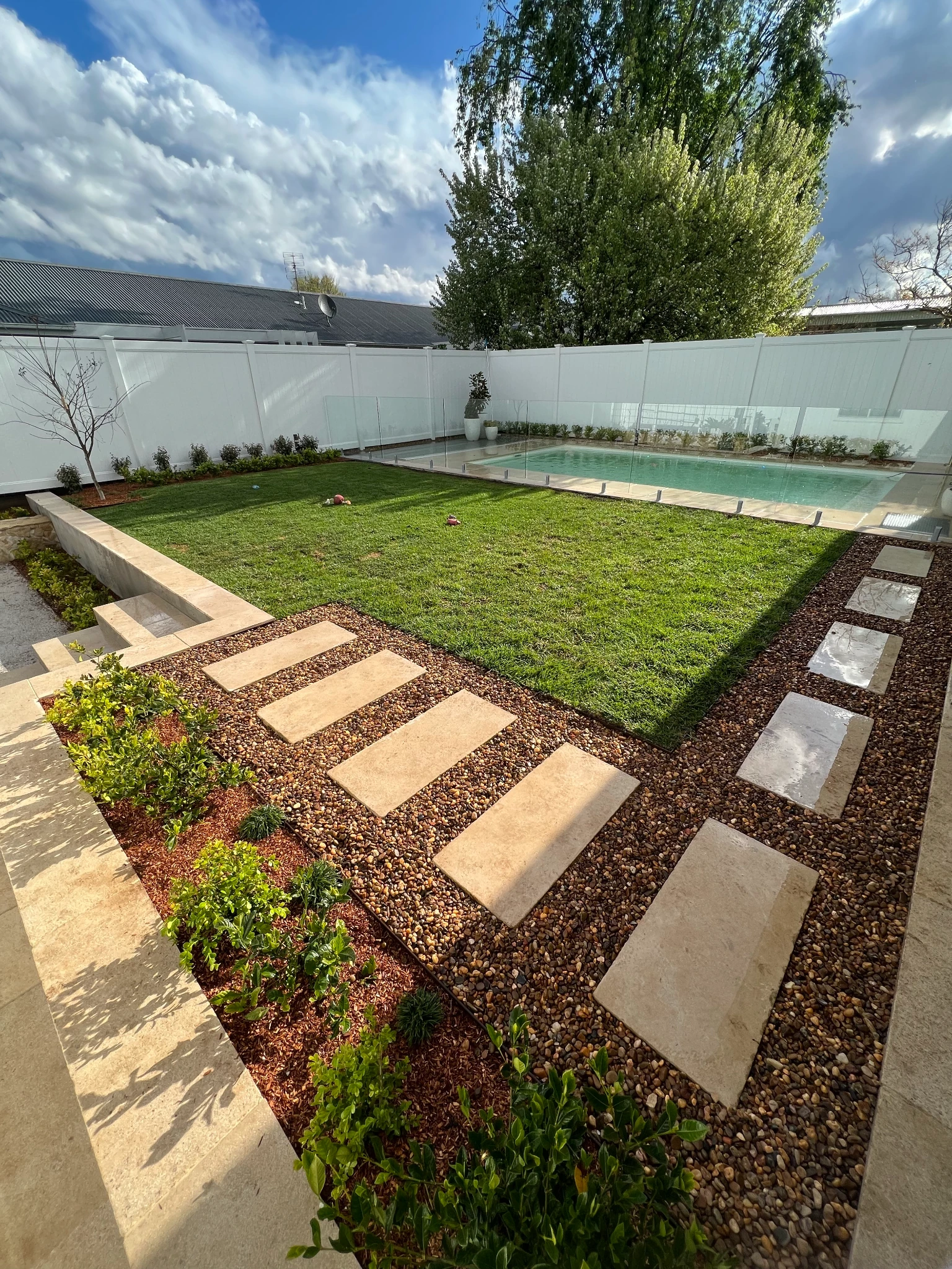 Stone stepping paths, green lawn and planted borders beside a glass-fenced swimming pool
